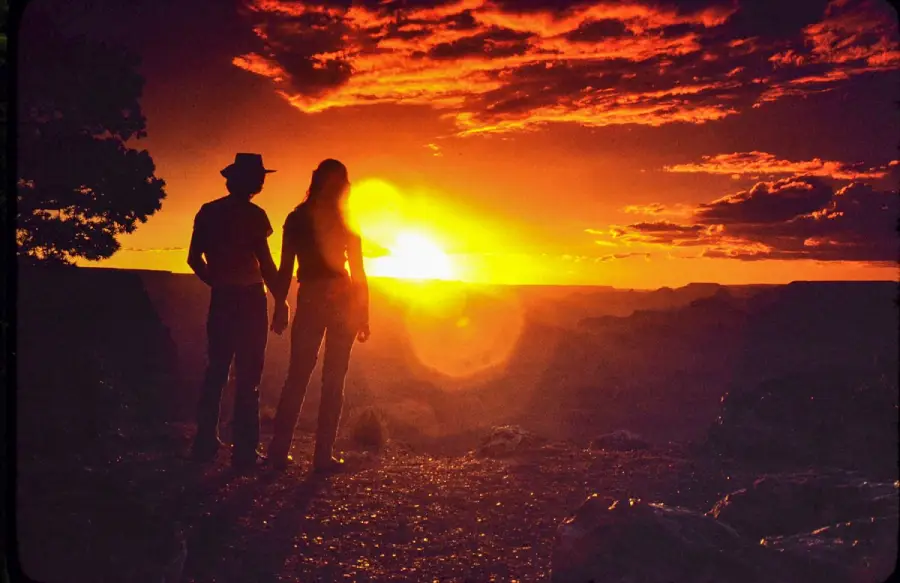 Two people stand hand in hand, silhouetted against a dramatic orange and yellow sunset sky, with clouds and distant canyon landscape visible in the background.