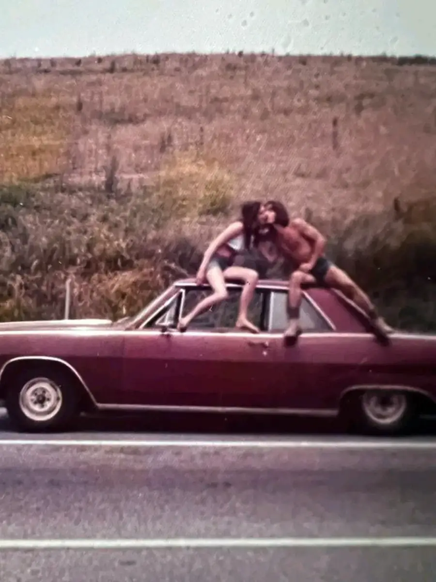 Two people in swimwear are sitting and playfully interacting on the roof and hood of a vintage red car parked by the roadside, with grassy hills in the background.