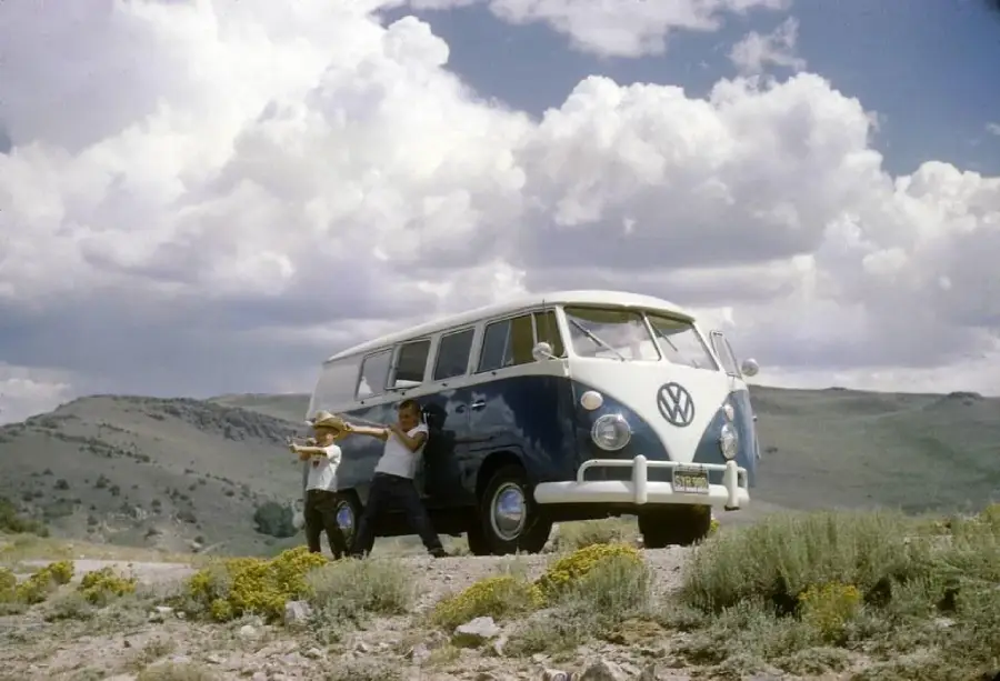 Two people stand beside a blue and white vintage Volkswagen van parked on a dirt road in a hilly landscape, with dramatic clouds in the sky overhead. Wild shrubs dot the foreground.