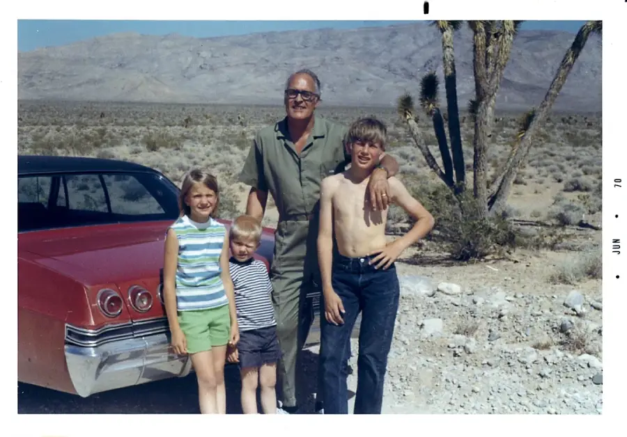 A man stands in a desert landscape with three children beside a vintage red car; one boy is shirtless, another is young and blonde, and a girl wears a striped shirt and green shorts. Distant mountains and a Joshua tree are visible.