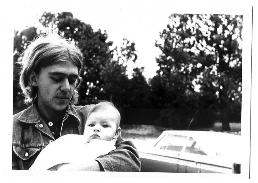 Black-and-white photo of a young man with long hair holding a baby outdoors, with trees and a parked car visible in the background. The man looks down while the baby looks toward the camera.