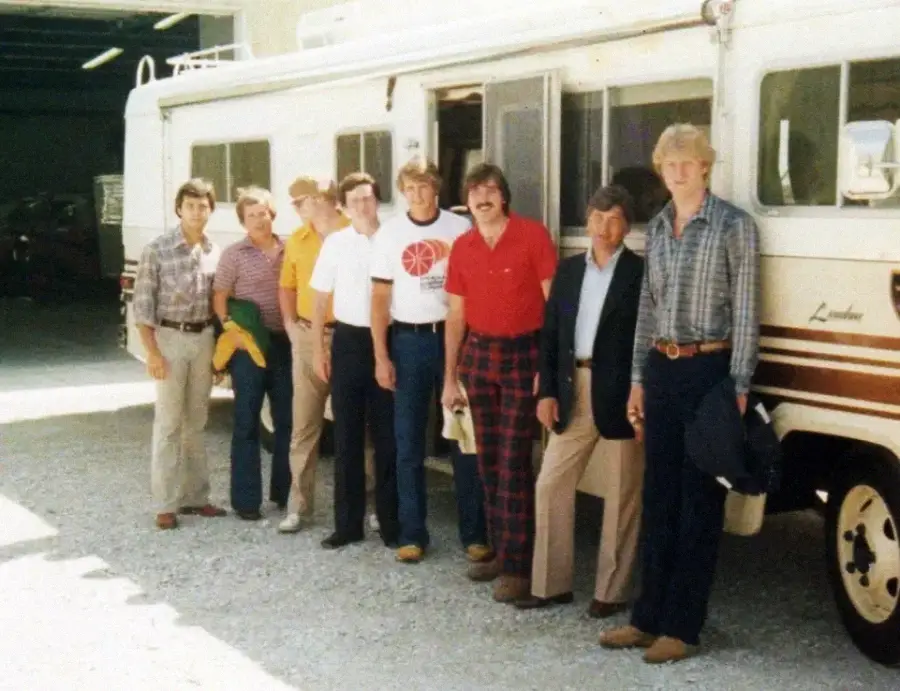 Eight men stand in a row outside a white recreational vehicle, posing for a group photo. Most are casually dressed in shirts and pants. The RV is parked on a gravel surface near a large building.