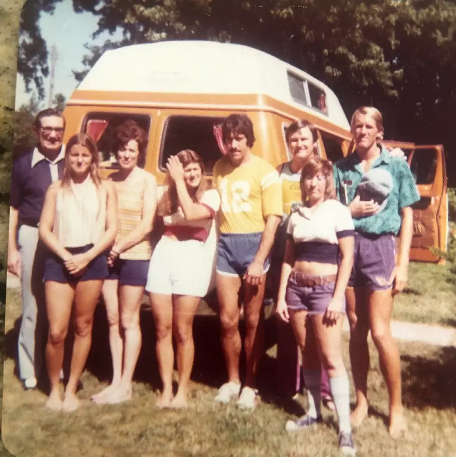 A group of seven adults stand smiling in front of an orange and white camper van on a sunny day. They are dressed in casual summer clothing, with trees visible in the background.
