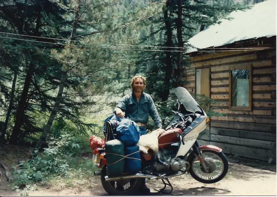 A person stands smiling next to a packed motorcycle in front of a wooden cabin, surrounded by tall trees and greenery on a sunny day.