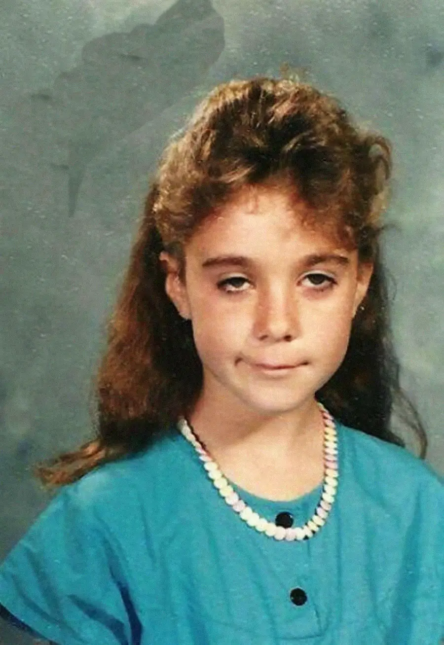 A young girl with long brown hair wearing a blue shirt and a candy necklace poses for a school photo, looking at the camera with a neutral, slightly unenthusiastic expression, against a gray mottled background.