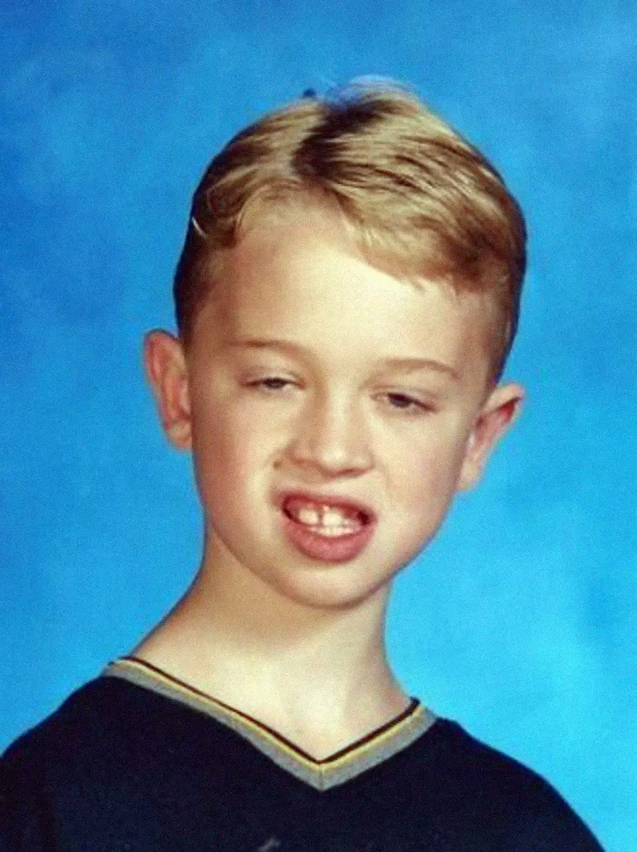 A young boy with short blonde hair and a dark shirt poses in front of a blue background, making an awkward facial expression with his mouth slightly open and eyes partially closed.