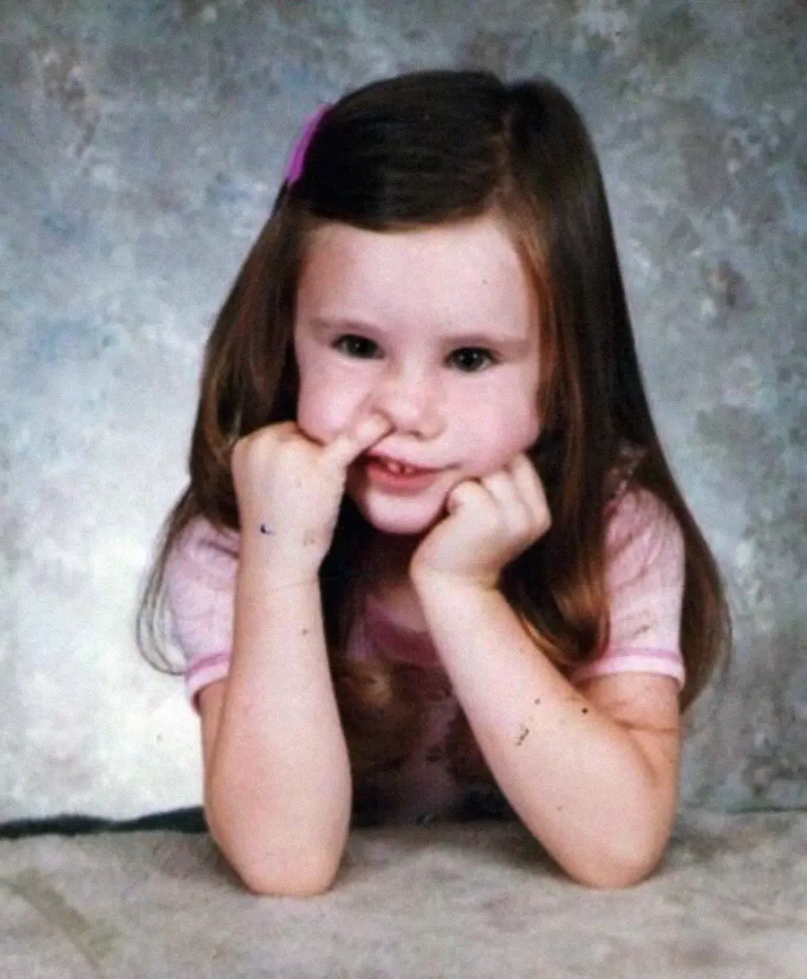 A young girl with long brown hair, wearing a pink shirt, smiles playfully at the camera while picking her nose with one finger, resting her head on her other hand.
