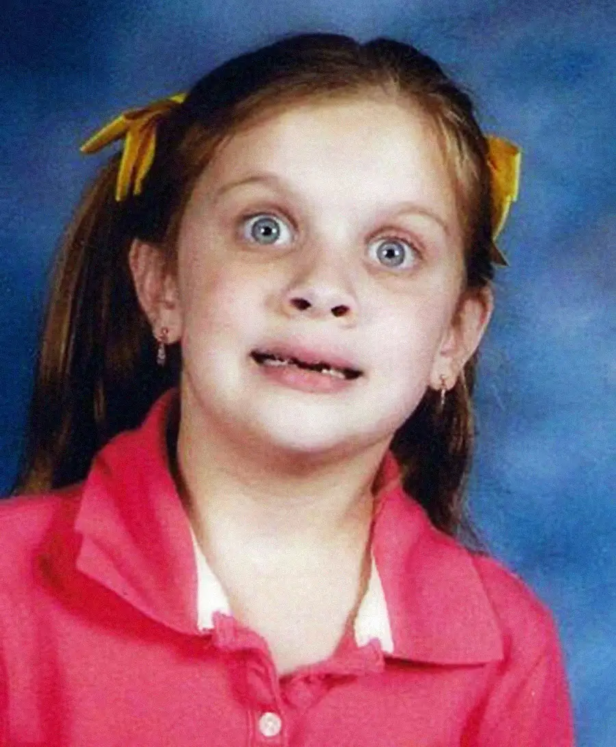 A young girl with wide blue eyes and a surprised expression, wearing a pink collared shirt and yellow bows in her brown hair, poses in front of a blue background for a school photo.