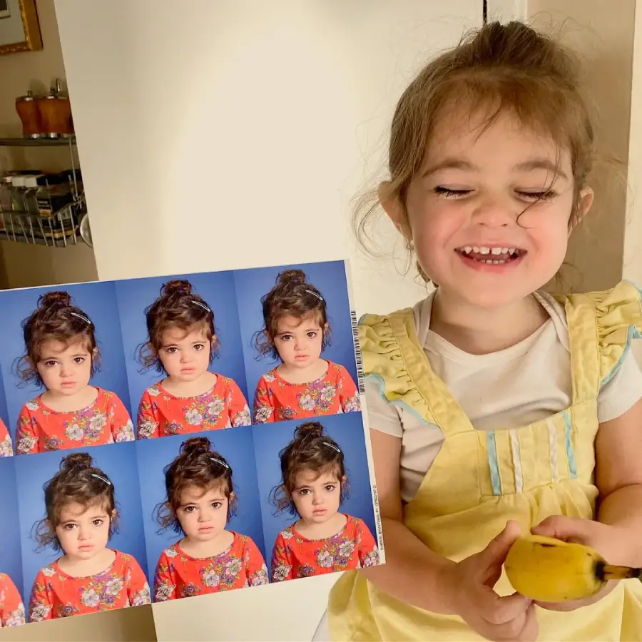 A young girl, smiling and holding a banana, stands next to a sheet of passport photos showing her with a serious expression in each picture. She wears a yellow dress over a white shirt.
