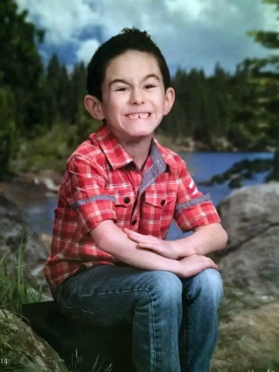 A young boy with dark hair, wearing a red plaid shirt and jeans, sits on a rock outdoors, smiling widely with arms crossed in front of a backdrop of trees and a lake.