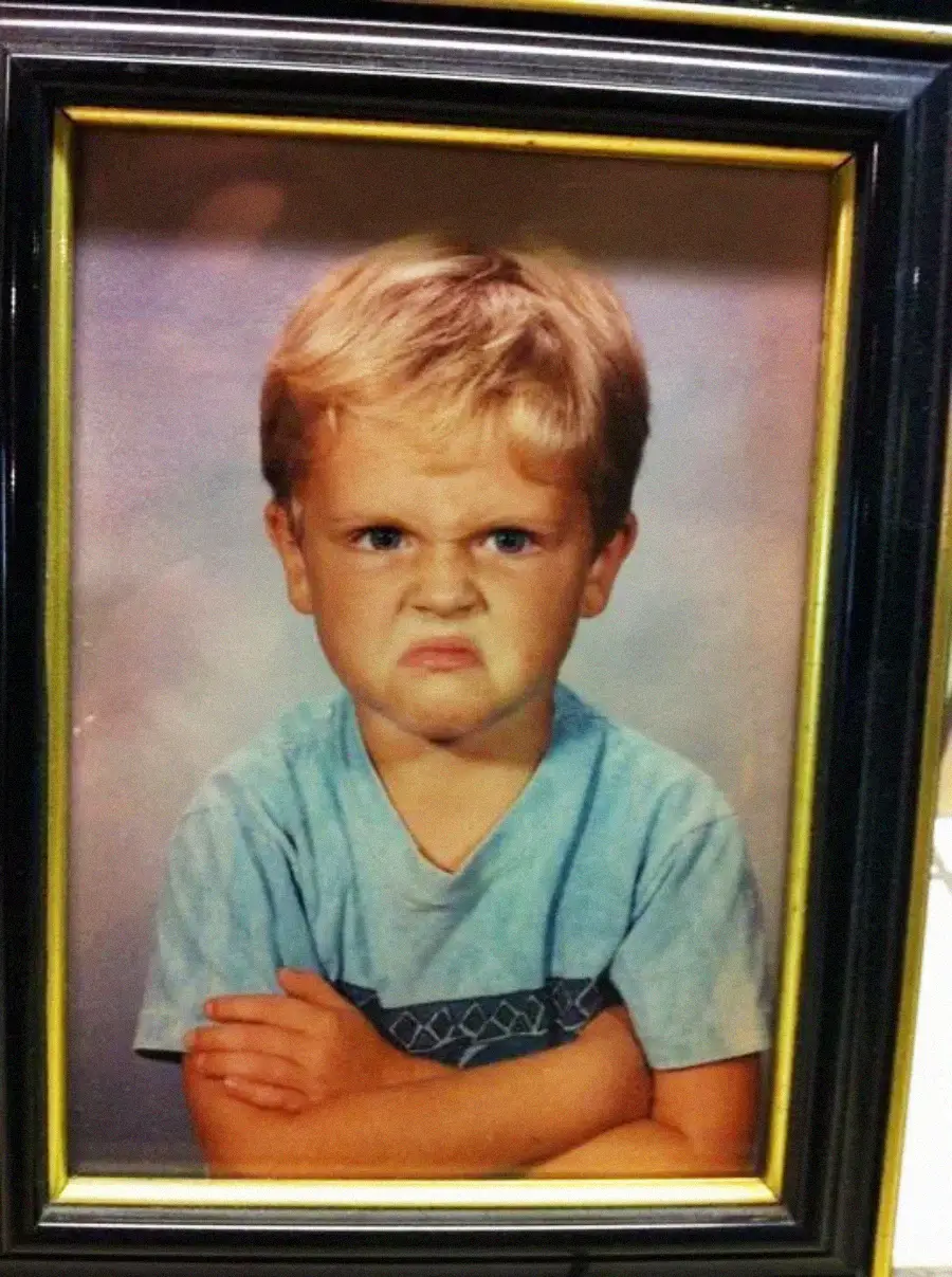 A young boy with blond hair and a blue t-shirt sits with his arms crossed, frowning and looking upset, in a framed portrait with a plain background.