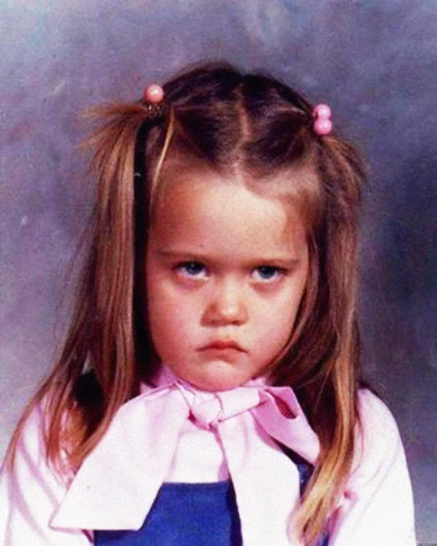 A young girl with long brown hair in pigtails and pink bows is wearing a pink shirt with a scarf and blue overalls, staring forward with an angry, unhappy expression against a gray backdrop.