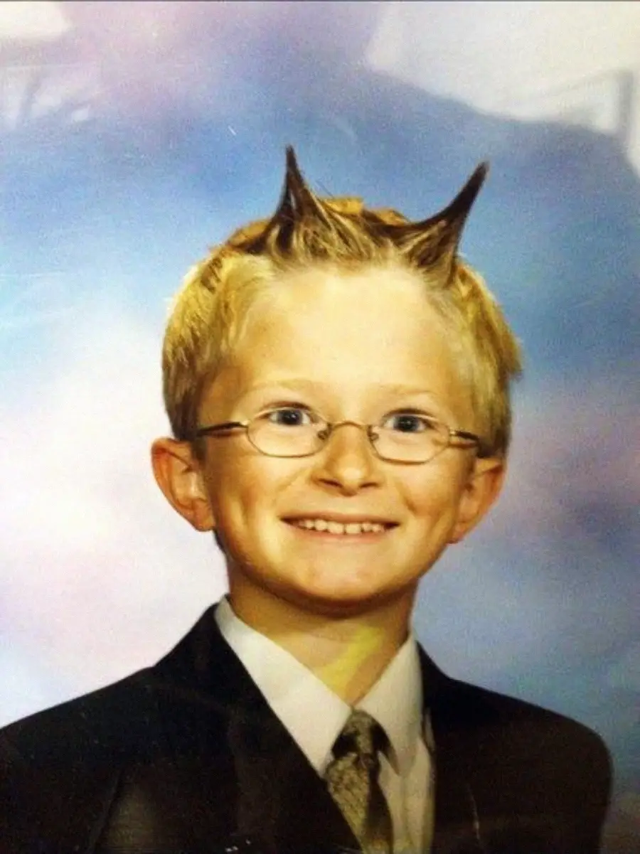 A young boy with glasses and a suit smiles at the camera. His blond hair is styled into two pointed horns on top of his head, giving a playful and mischievous look. The background is softly blurred pastel colors.