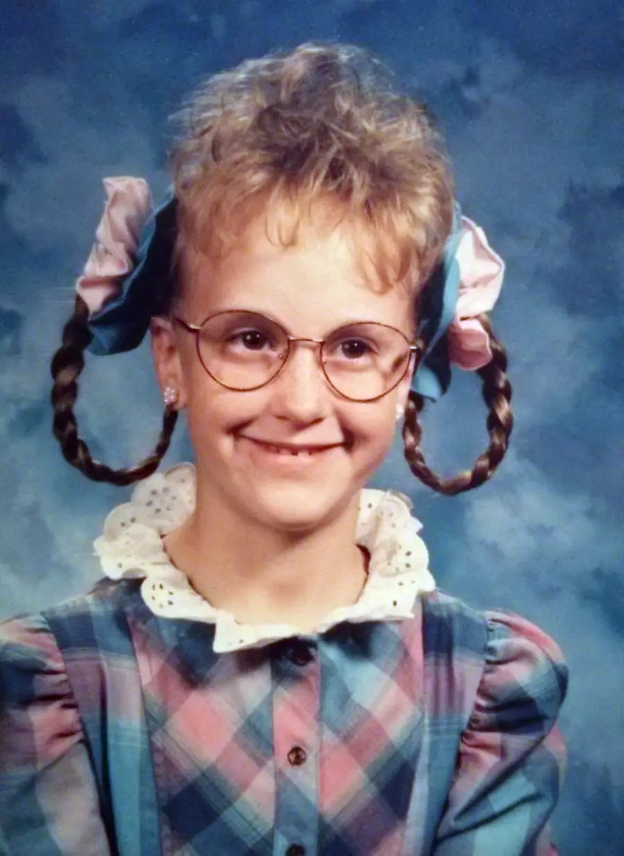 A young girl with glasses and braided pigtails sticking out sideways, tied with large pink and blue ribbons, smiles in a school portrait. She wears a plaid dress with puffed sleeves and a white lace collar.