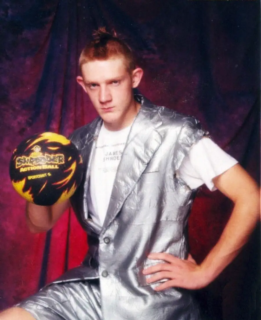 A young man in a silver metallic suit poses with a yellow and black bowling ball against a dark, textured backdrop. He has short, spiked hair and a serious expression, with one hand on his hip.