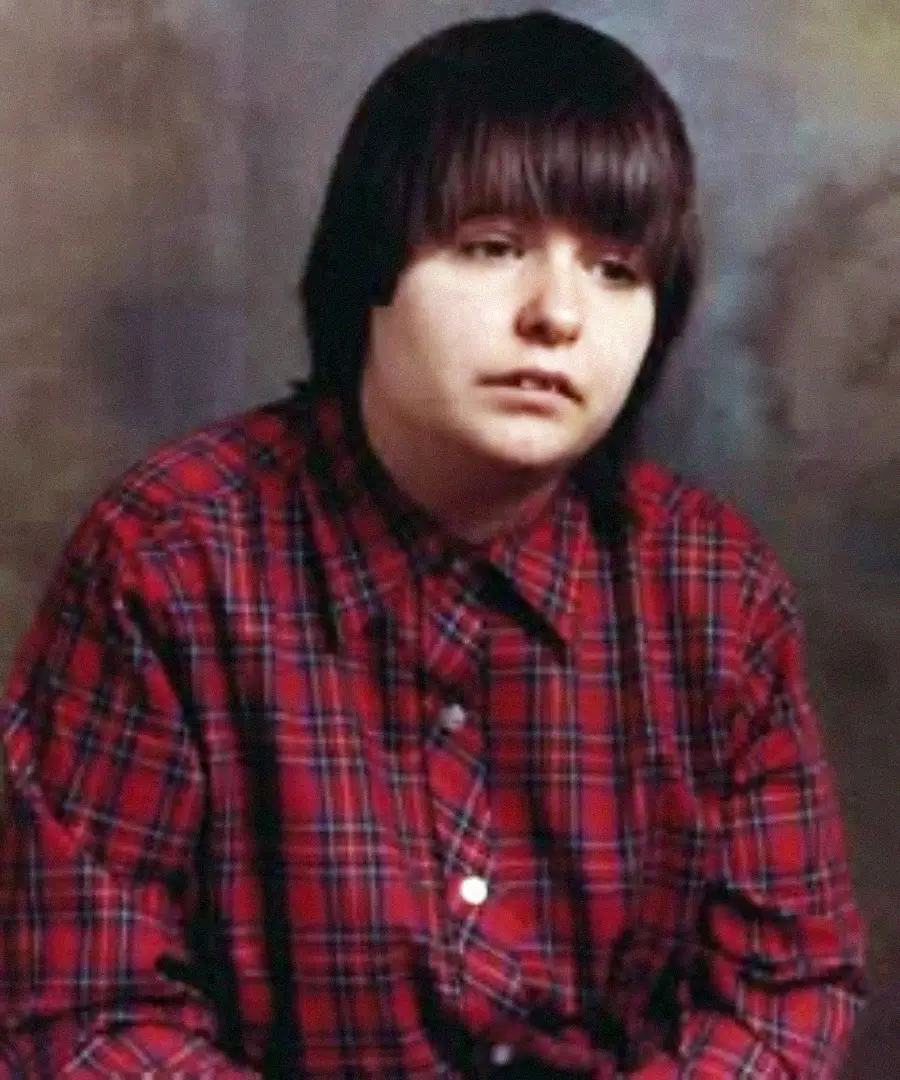 A young person with straight, dark hair and bangs sits in front of a neutral, blurred background. They are wearing a red and black plaid button-up shirt and have a neutral or slightly pensive expression.