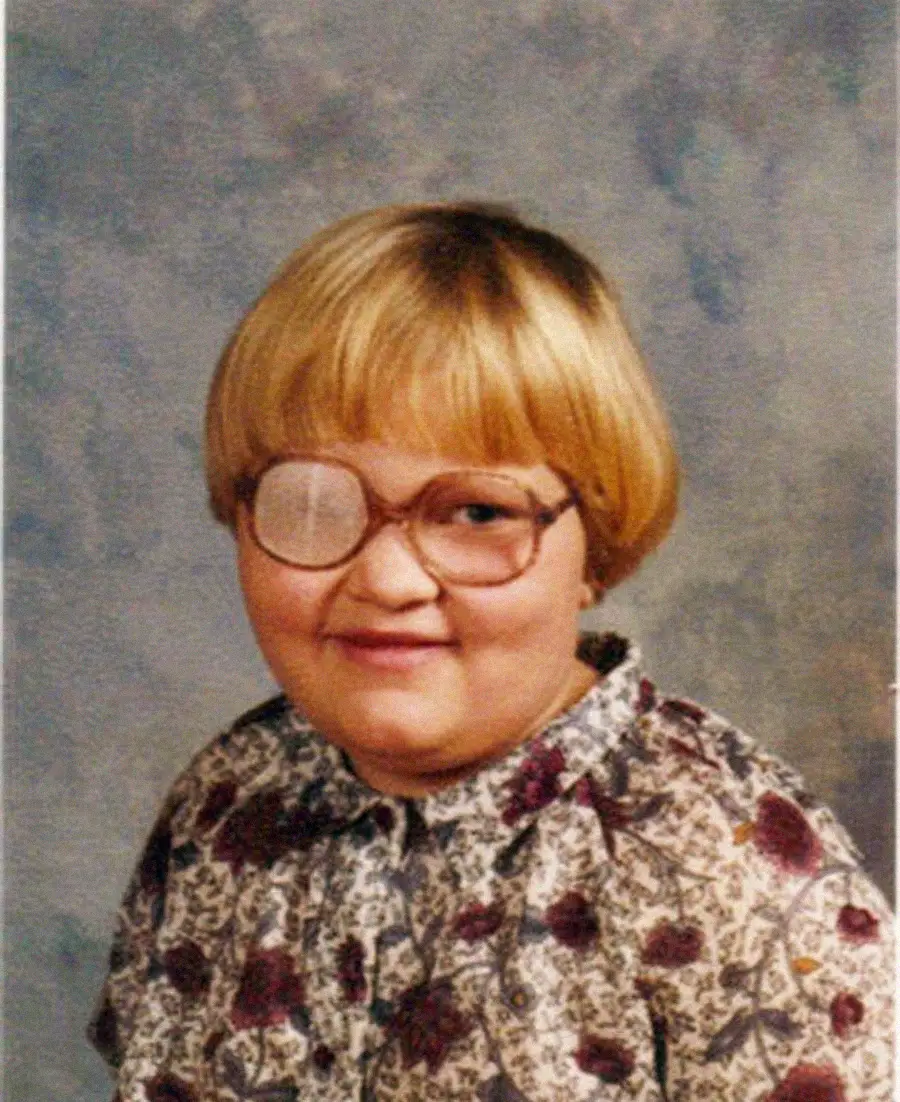 A child with short blond hair and glasses wears a patterned blouse and a white eye patch over the left eye, smiling in a school portrait against a mottled gray background.