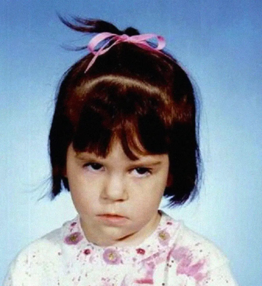 A young girl with short brown hair and bangs wears a pink and blue bow, a white shirt with pink patterns, and looks directly at the camera with a frown against a plain blue background.