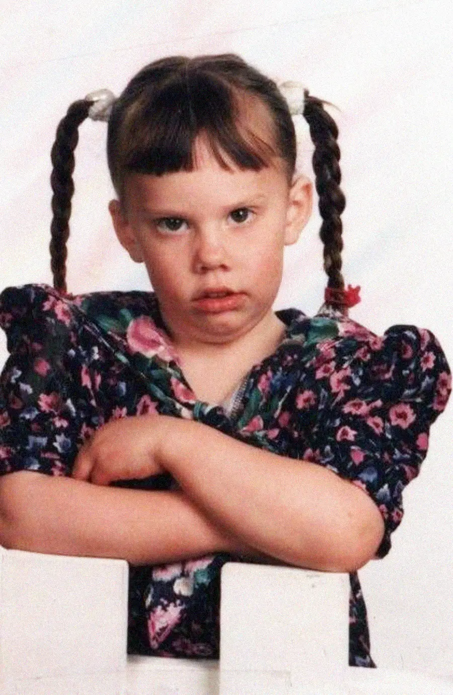 A young girl with braided pigtails and bangs, wearing a floral dress, stands with her arms crossed and an unhappy or upset expression on her face against a plain light background.