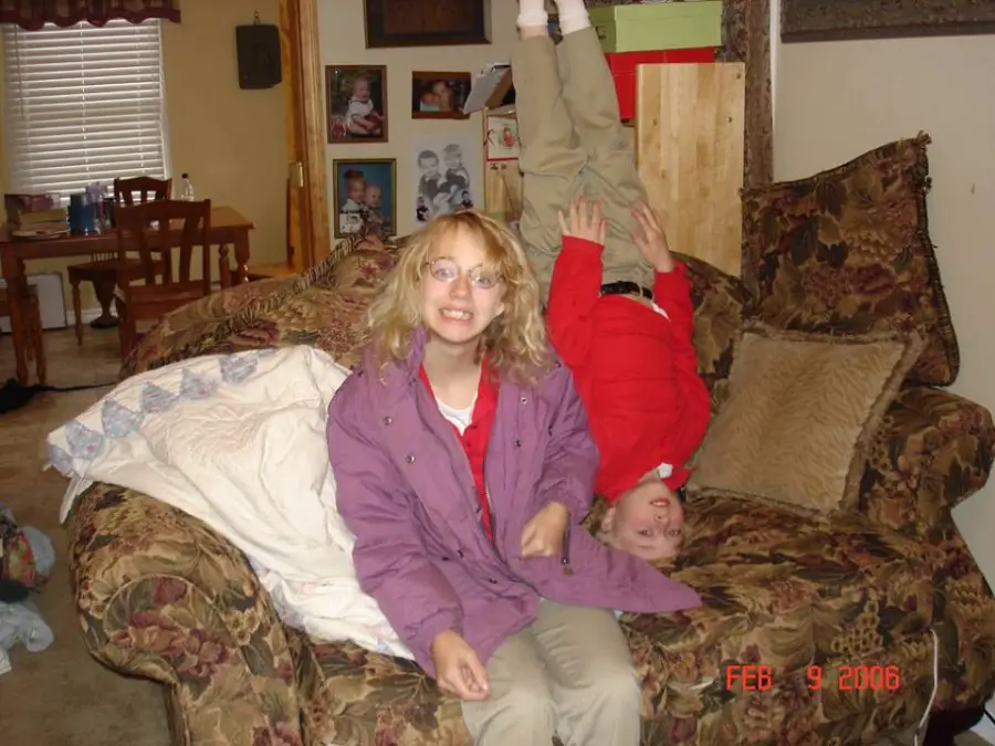 A girl in glasses and a purple jacket smiles on a patterned couch while a boy in red and khaki does a headstand beside her. The cozy living room has family photos on the wall and a quilt draped on the couch.