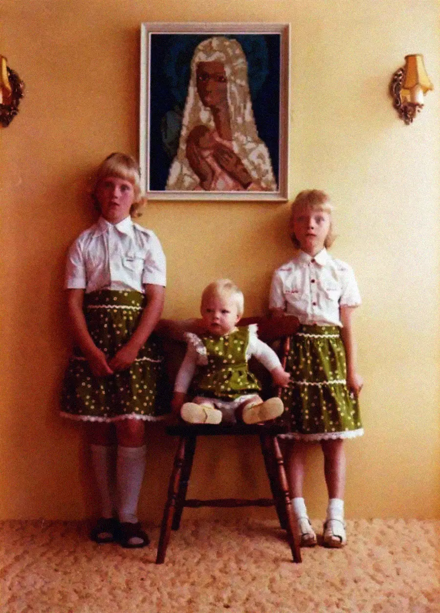 Three young children pose indoors against a yellow wall, with two girls standing in matching green dresses and a baby sitting in a chair between them. A framed portrait and two sconces hang on the wall above them.