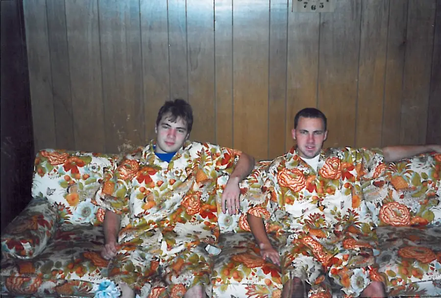 Two young men sit on a sofa, both wearing clothes made from the same orange floral fabric as the couch and pillows, blending into the patterned background against a wood-paneled wall.