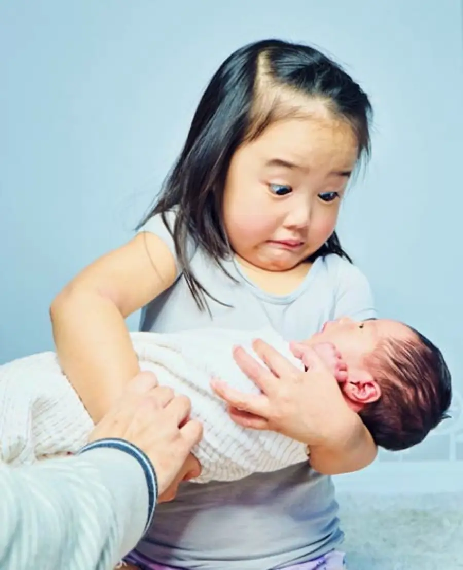 A young girl with a surprised expression holds a newborn baby wrapped in a white blanket while an adult hand supports the baby.