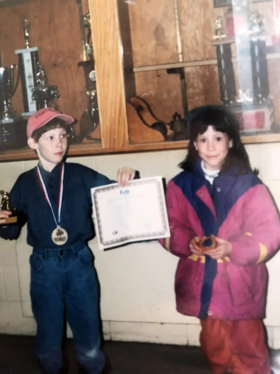 Two children stand indoors in front of a trophy case. The boy on the left wears a medal and cap, holding a trophy and certificate. The girl on the right wears a pink jacket and holds a small object, both with neutral expressions.