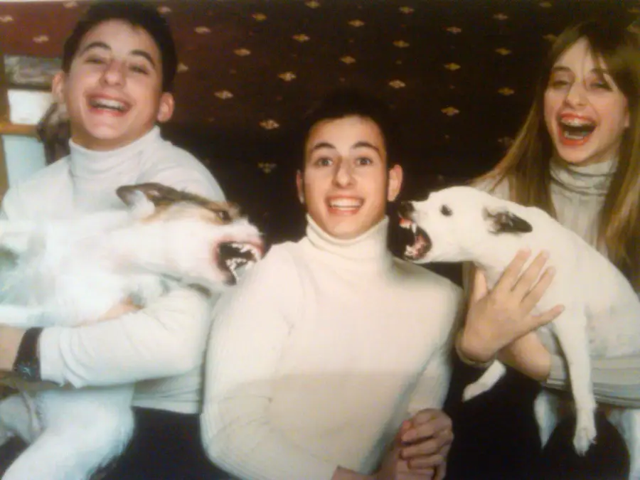 Three people in white turtlenecks smile widely while holding two dogs that appear to be playfully snarling at each other, creating a humorous and lively scene.