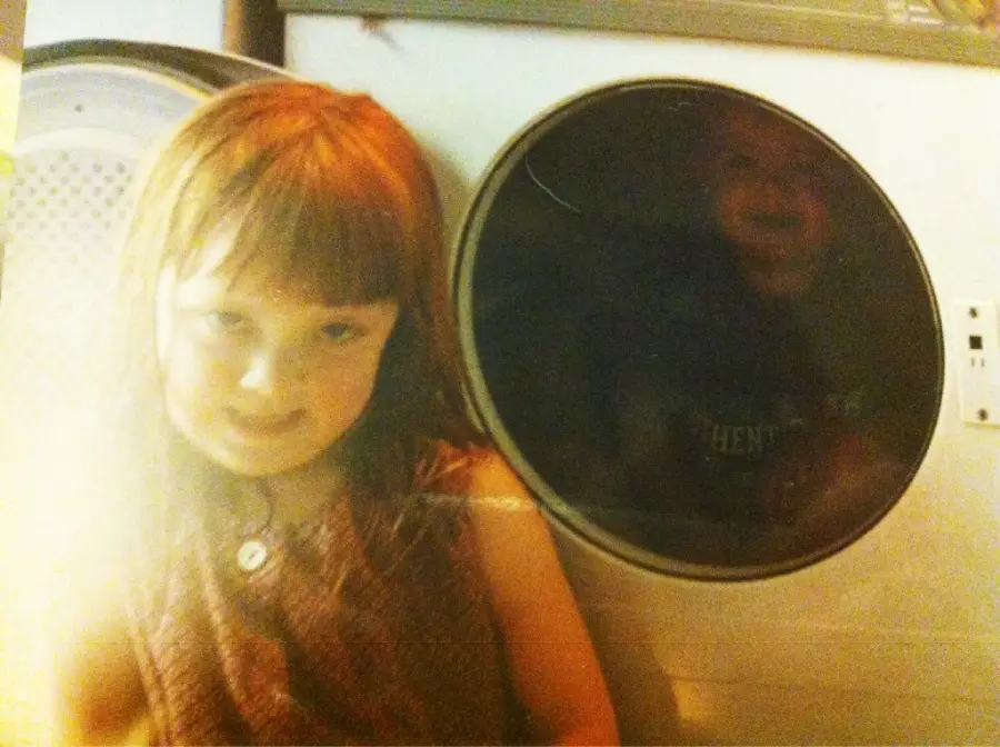 A young girl with reddish-brown hair sits in front of a dryer, smiling slightly. Inside the dryer, a faint reflection or image of another child is visible through the round glass door.