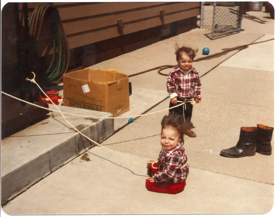 Two young children with static, messy hair are playing outside. One is standing, the other is sitting on the ground near a cardboard box, ropes, a pair of boots, and a garden hose. Both wear plaid shirts and red pants.
