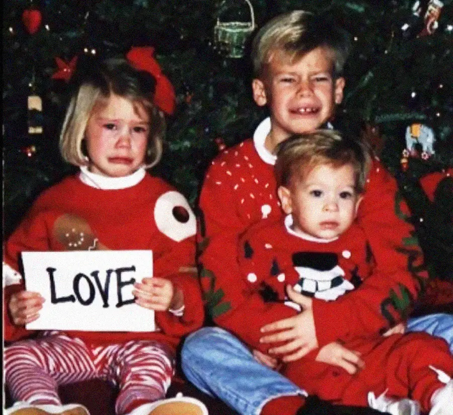 Three young children sit in front of a Christmas tree. The girl on the left, holding a "LOVE" sign, looks upset and tearful. The boy in the middle is squinting and grimacing, while the toddler on his lap looks calm.