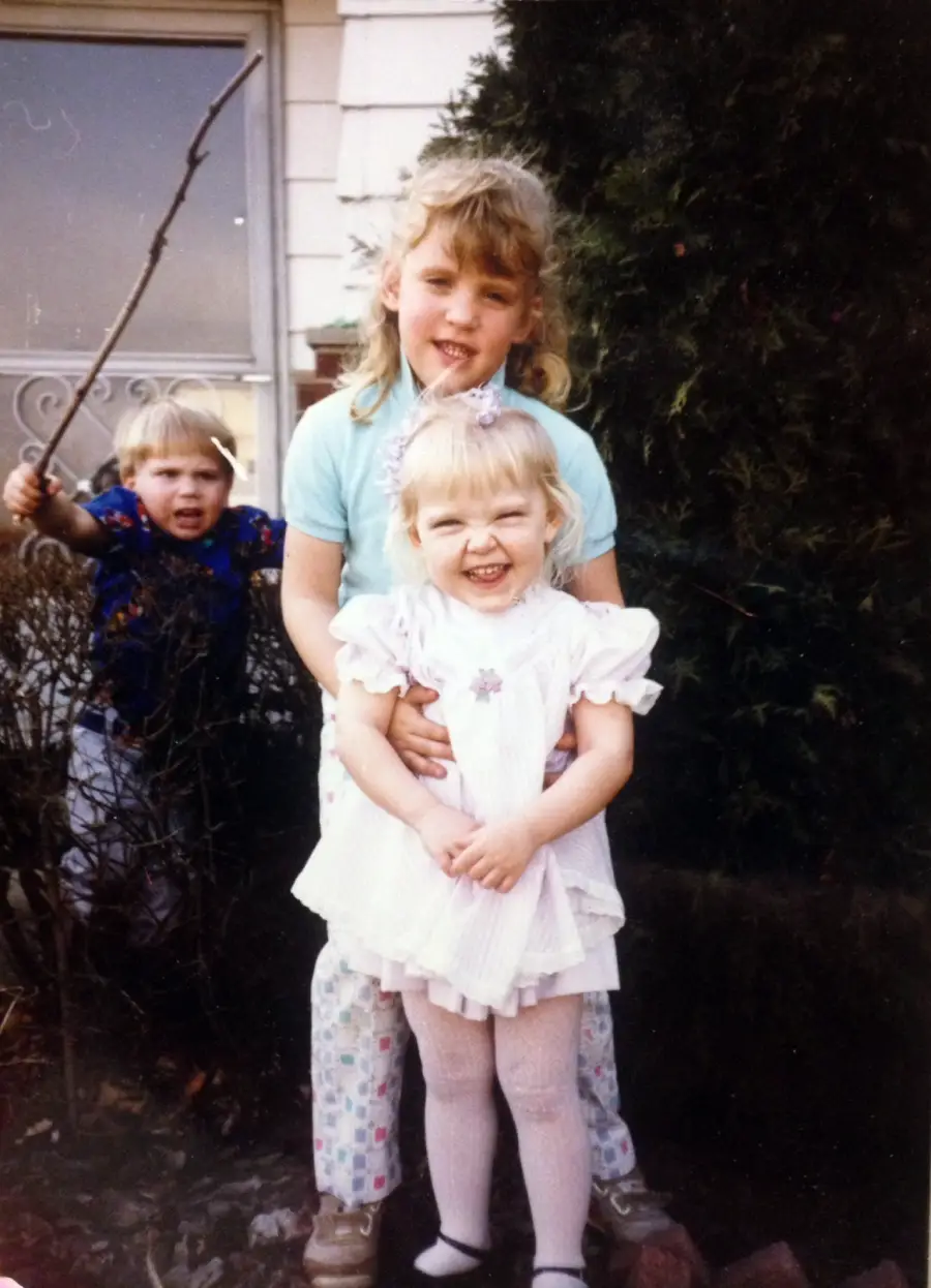 Three young children pose outside a house. Two girls stand in front, smiling, with the older girl hugging the younger. A boy stands behind them in the bushes, holding a stick and making an expressive face.