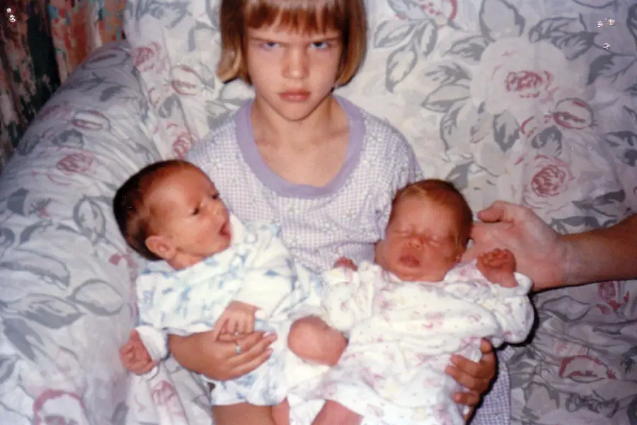 A young girl with a serious, unhappy expression sits on a floral couch, holding two newborn babies wrapped in blankets, one in each arm. An adult hand supports one of the babies.