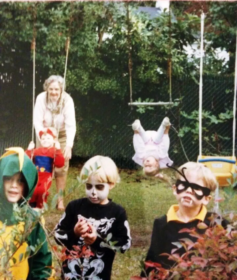 Four young children in costumes and face paint stand in a yard, with one child on a swing upside down and an elderly woman smiling behind them. The scene is playful and lively, surrounded by greenery.