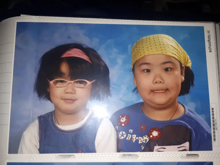 Two young children pose in front of a blue backdrop. The child on the left wears pink glasses, a pink headband, and a denim dress. The child on the right wears a yellow bandana and blue shirt, smiling with a playful expression.