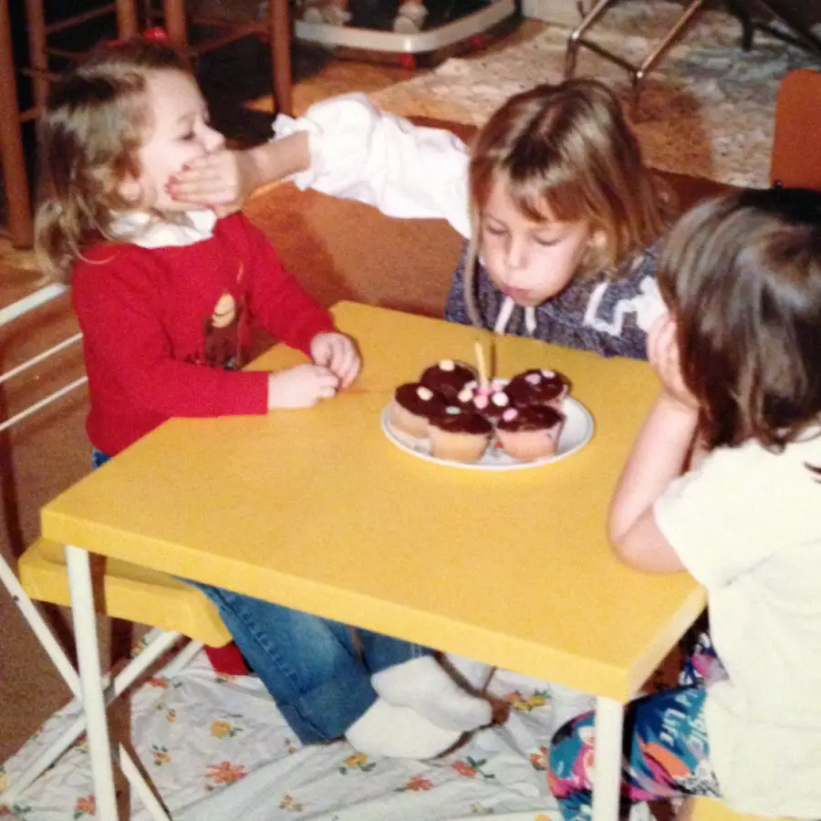 Three young children sit at a small yellow table with cupcakes. One child blows out candles while another covers the mouth of the child beside her. The third child watches with hands on her cheeks.