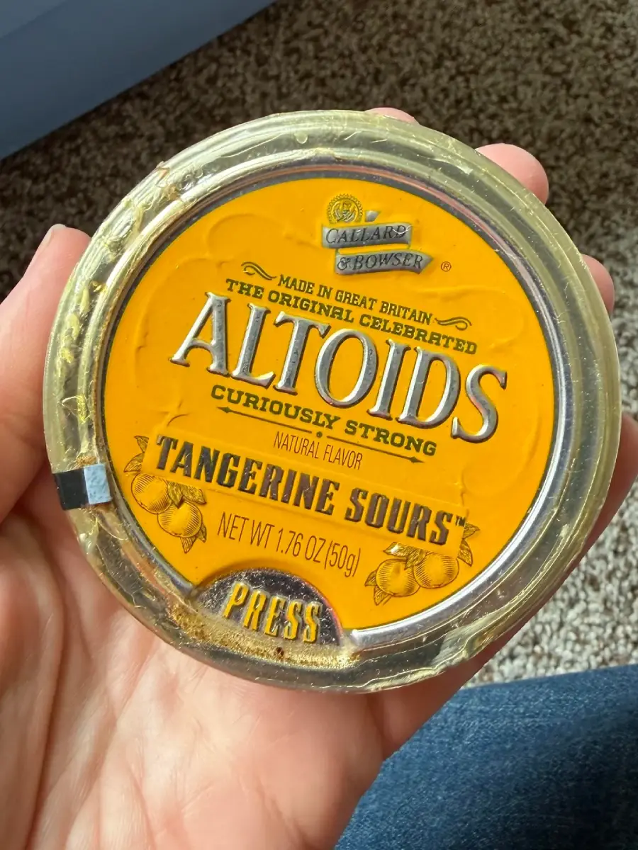 A hand holds a round, orange Altoids tin labeled "Tangerine Sours," with some wear and discoloration on the lid. The background includes carpet and part of a blue object.