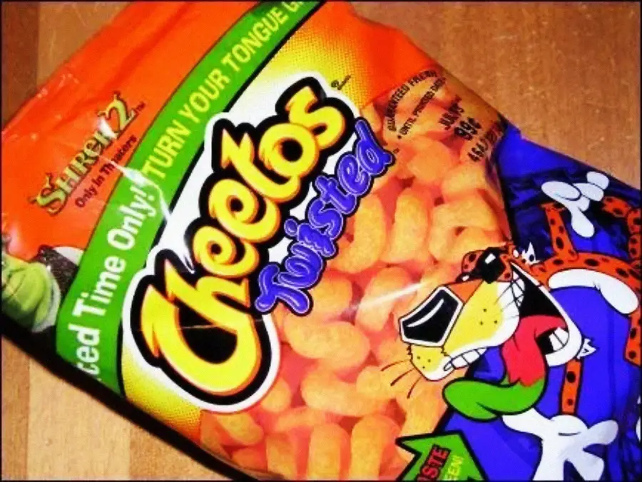 A brightly colored bag of Cheetos Twisted snacks sits on a wooden surface. The packaging features the Cheetos logo, mascot Chester Cheetah, and the phrase “Limited Time Only! Turn Your Tongue Green!”