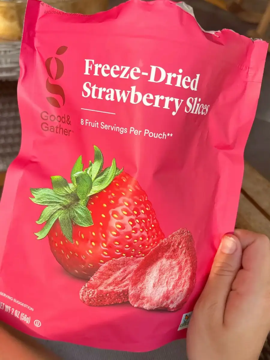A hand holds a pink bag of Good & Gather Freeze-Dried Strawberry Slices, featuring images of a fresh strawberry and freeze-dried strawberry pieces on the front. The bag indicates 8 fruit servings per pouch.