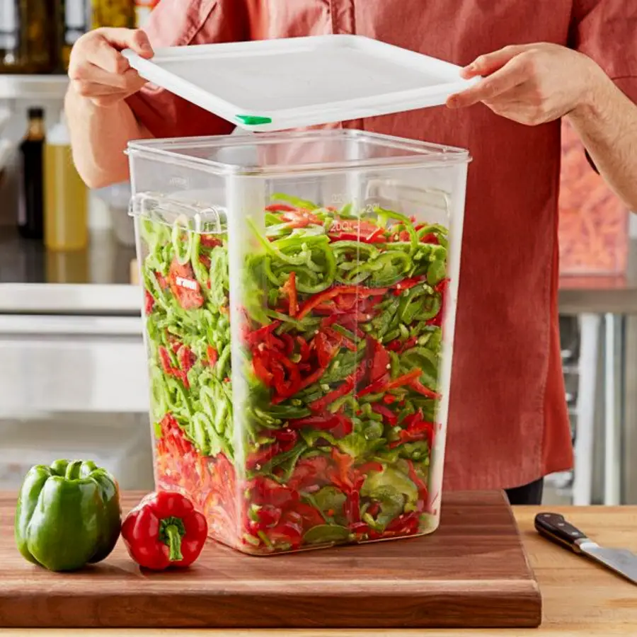 A person in a red shirt places a lid on a large clear container filled with sliced green and red bell peppers, with whole bell peppers on a wooden cutting board nearby.