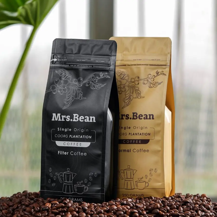Two coffee bags labeled "Mrs. Bean" stand side by side on a bed of coffee beans, with one black bag for filter coffee and one beige bag for normal coffee. A blurred green plant and window appear in the background.