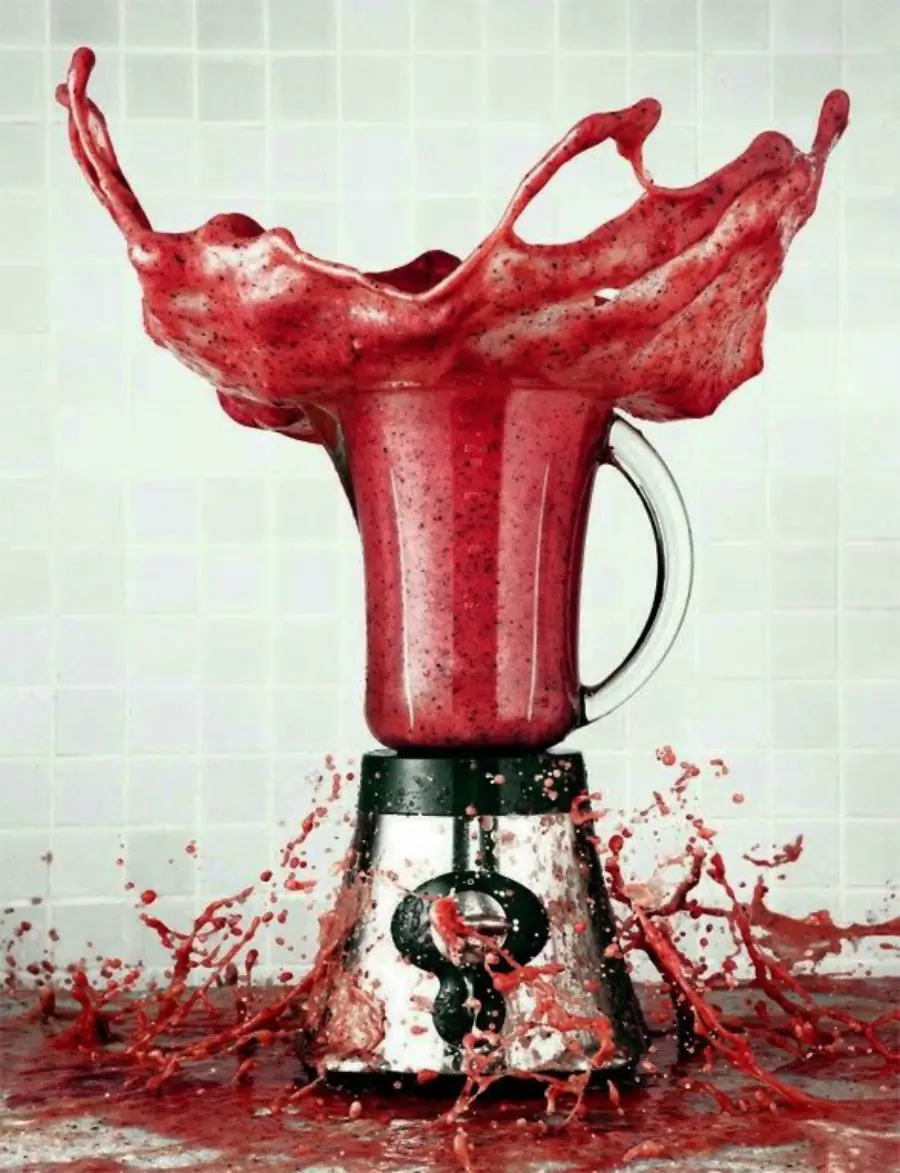 A blender filled with a red smoothie has its lid off, causing the liquid to splash dramatically out of the top and spill over the counter against a tiled background.