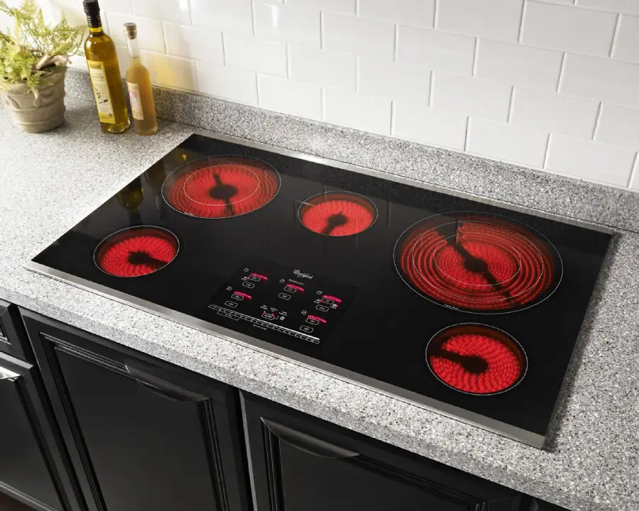 A black glass electric cooktop with five red-lit heating elements on a gray countertop, with white tile backsplash, olive oil bottles, and a small potted plant nearby.