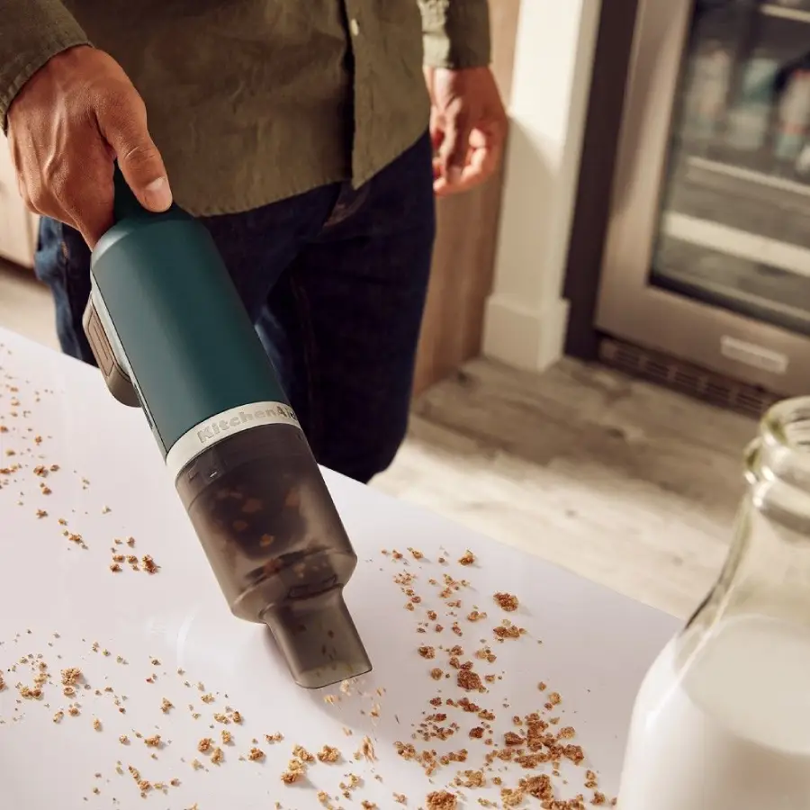 A person uses a handheld vacuum to clean up crumbs from a white surface near a spilled bottle of milk in a kitchen.