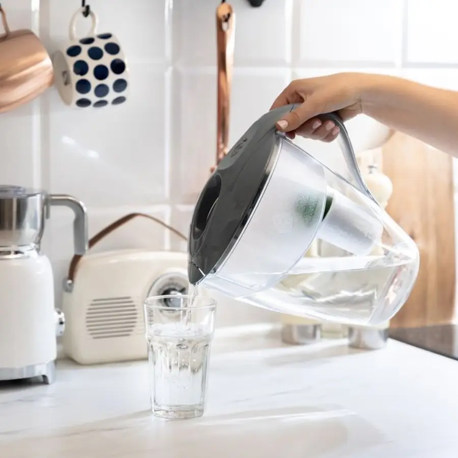 A hand pours water from a large, clear pitcher with a gray lid into a glass on a white kitchen counter; mugs and utensils hang on the tiled wall in the background.