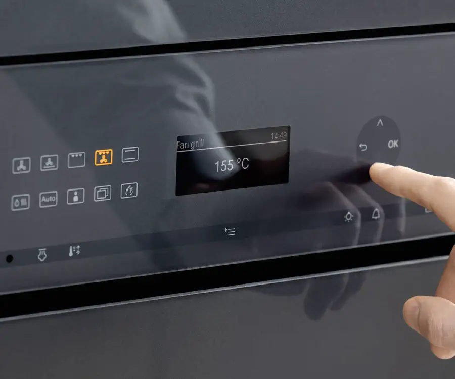 A hand is pressing the "OK" button on a modern oven's digital touchscreen control panel, which displays "Fan grill" and a temperature of 155°C.