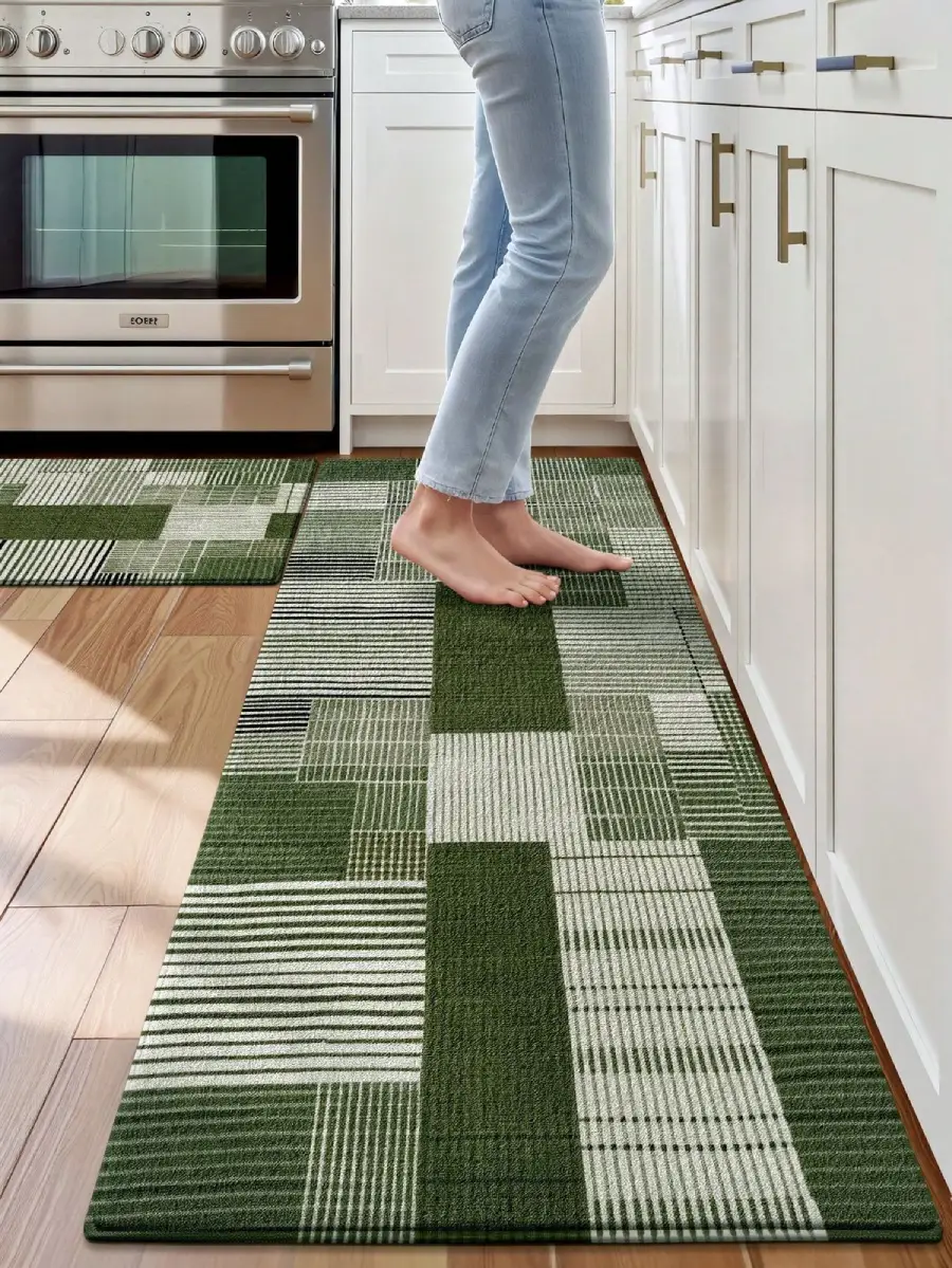 A person stands barefoot on a green and white geometric-patterned rug in a modern kitchen, next to white cabinets and a stainless steel oven.