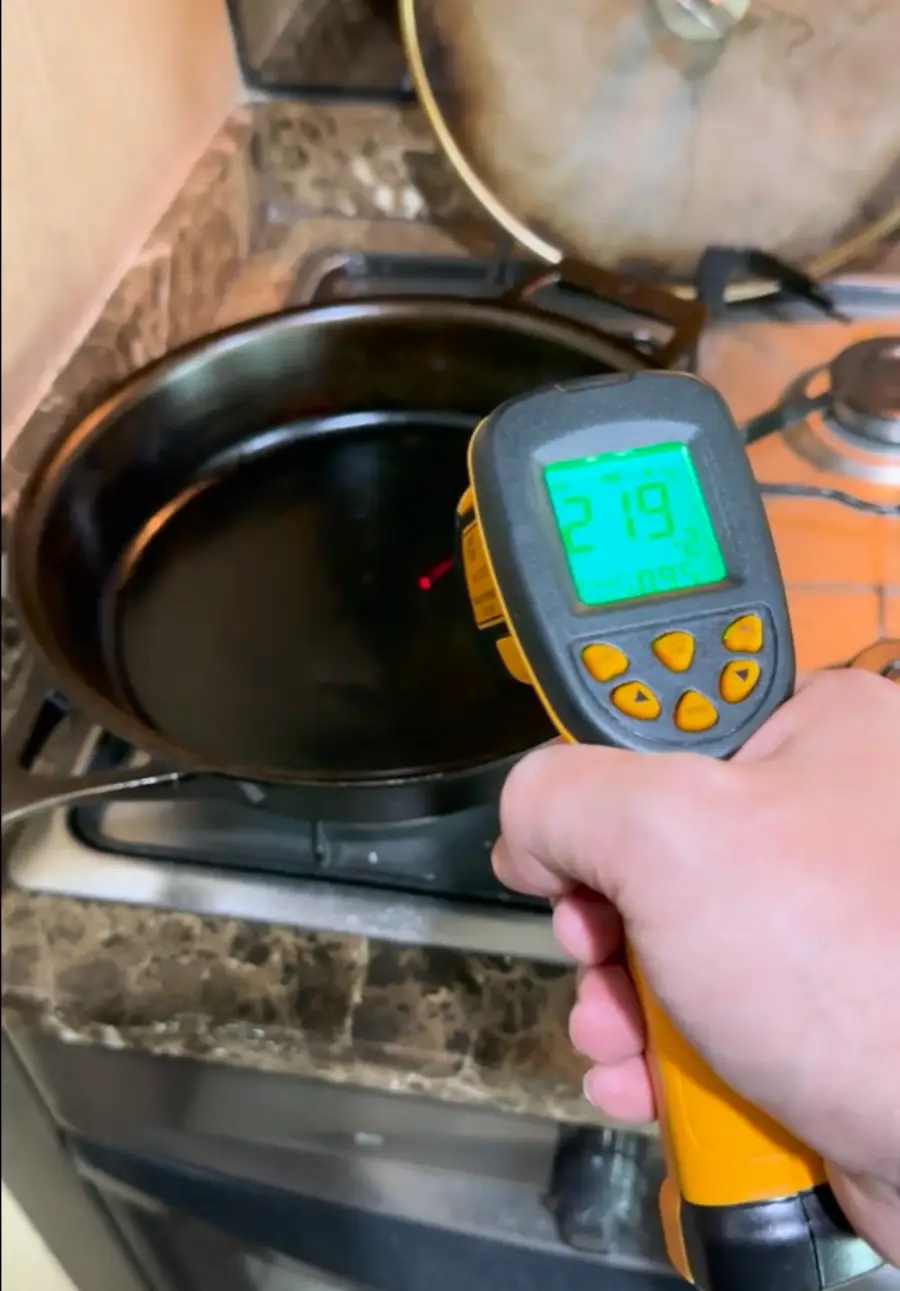 A hand holds an infrared thermometer pointed at a black frying pan on a stove. The thermometer display shows a temperature of 219 degrees. A pot with a glass lid sits on another burner in the background.