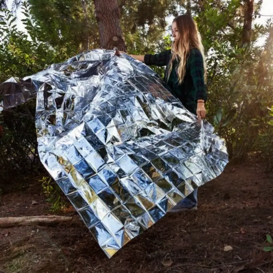 A person stands outdoors in a wooded area, holding and unfolding a large, shiny, silver emergency blanket that reflects sunlight.